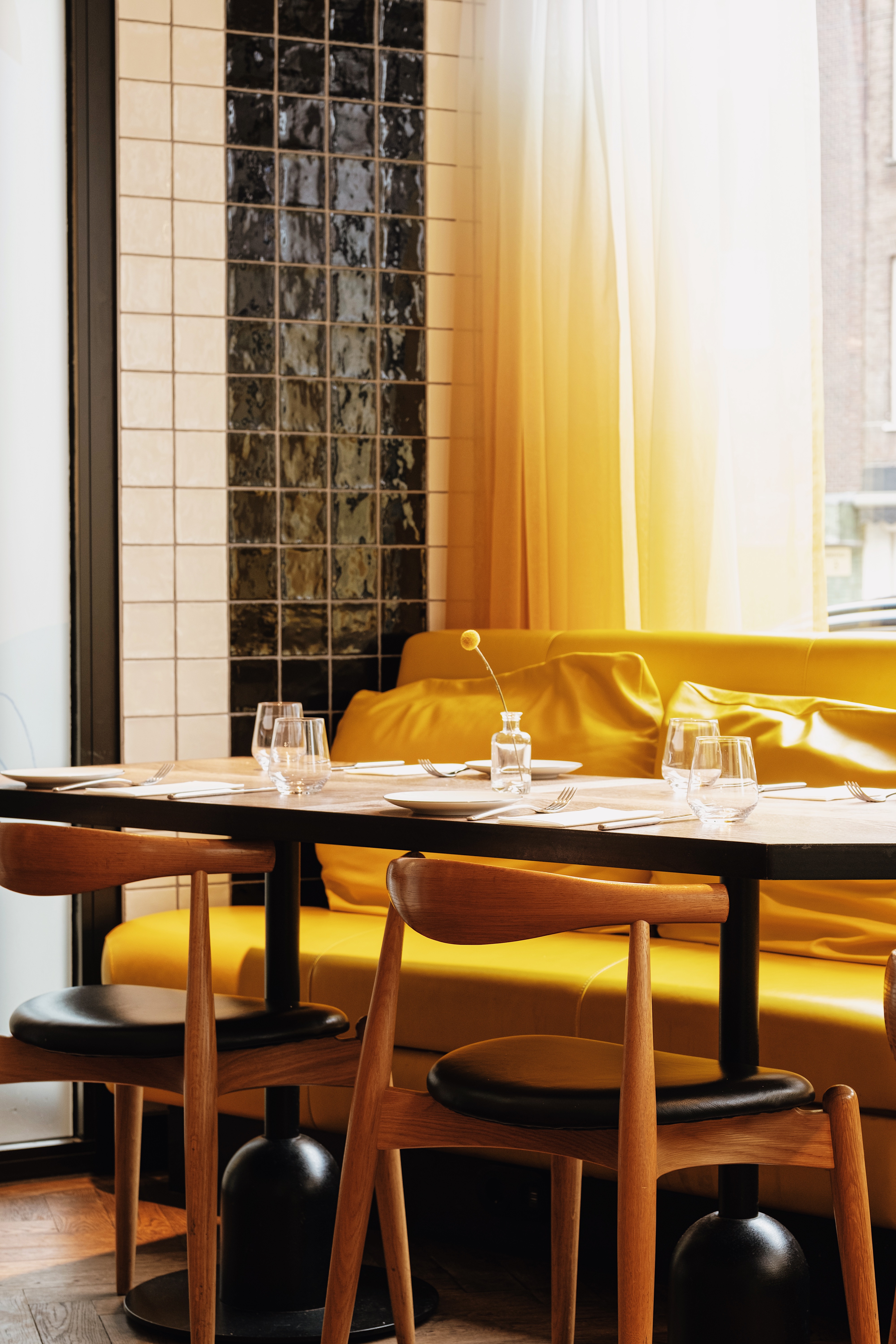 An intimate table setup with a yellow couch and cushions, glassware, and a small flower vase at Celia Amsterdam.