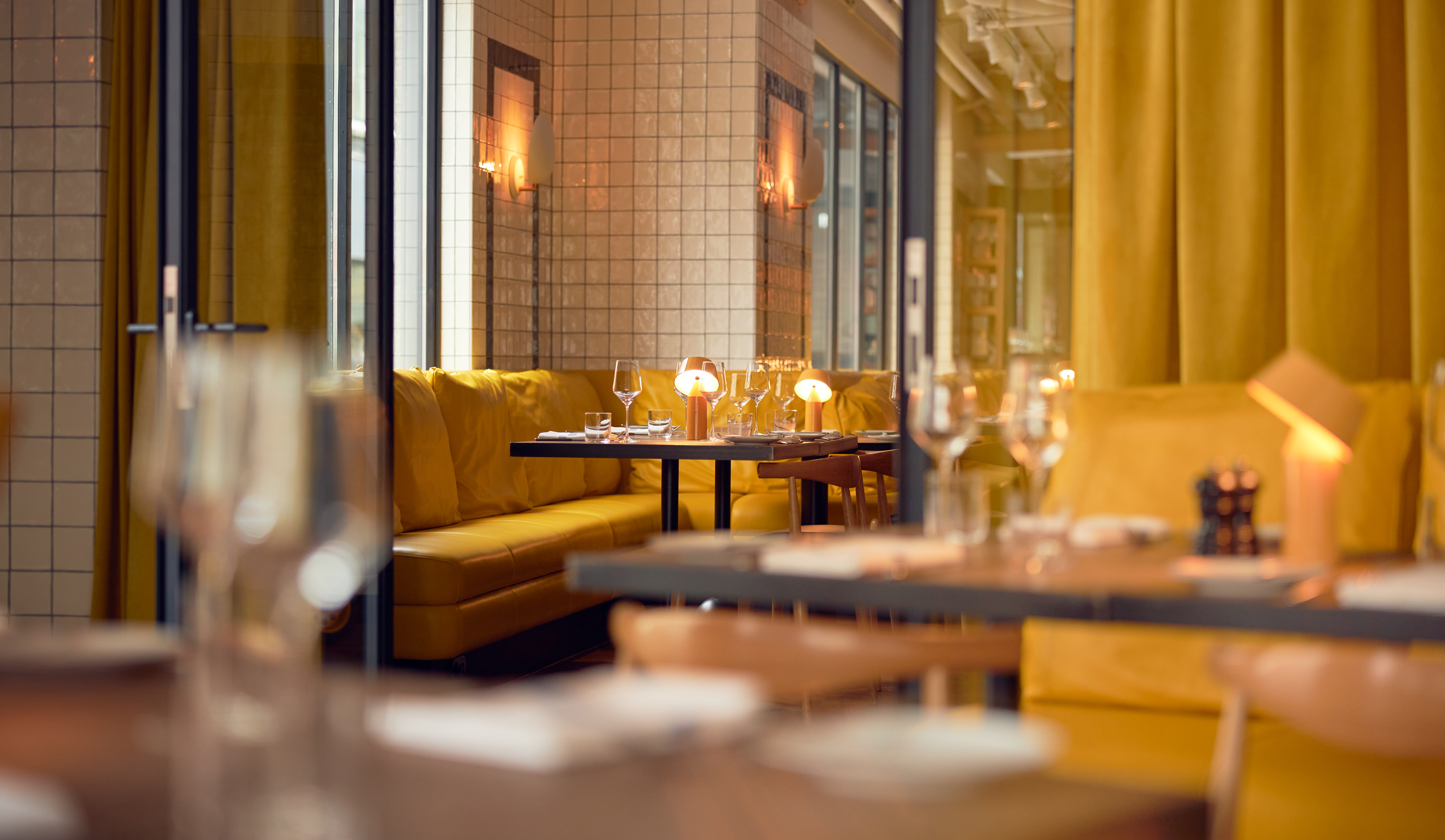 A cozy dining area with yellow cushions and leather couch, a table set for two, and soft lighting in Celia Amsterdam.