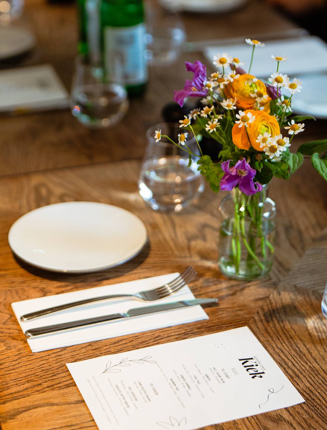 A wooden table is set at Celia with a menu and a vase of fresh flowers prepared for a private meal at an Amsterdam event.