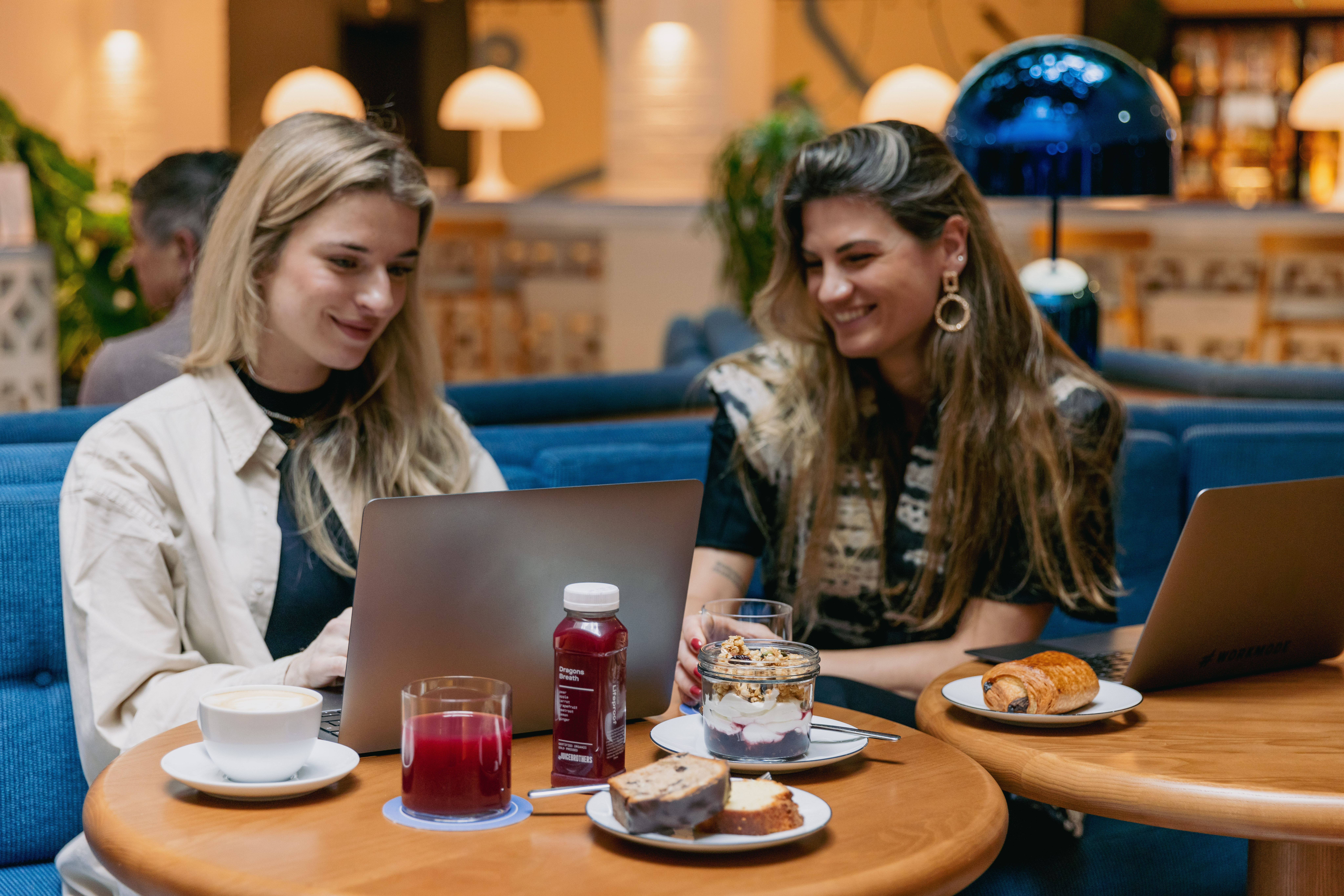 Two women enjoying sits by a laptop on a wooden table with coffee, juice, and dessert at Celia Amsterdam's co-working café.