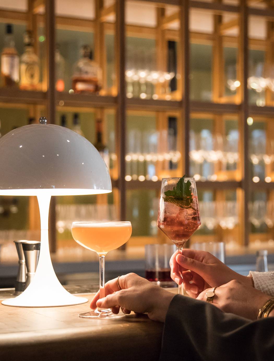 Clinking cocktail glasses in front of a well-stocked bar, with soft lighting casting a warm glow and a white lamp at Celia.