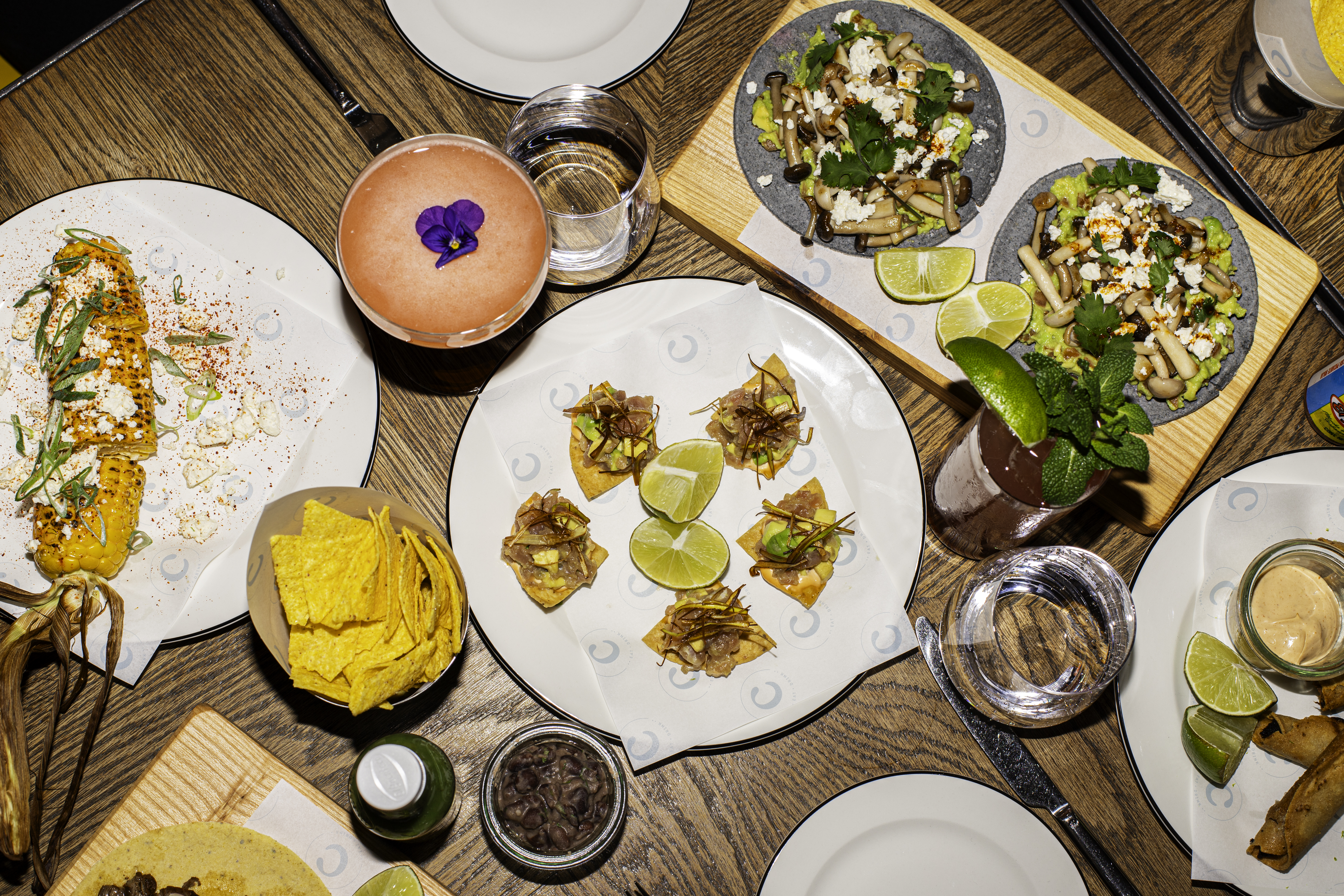 A variety of small bites, tacos, and cocktails on a table with lime wedges at Taco Tuesday at Celia in Amsterdam.