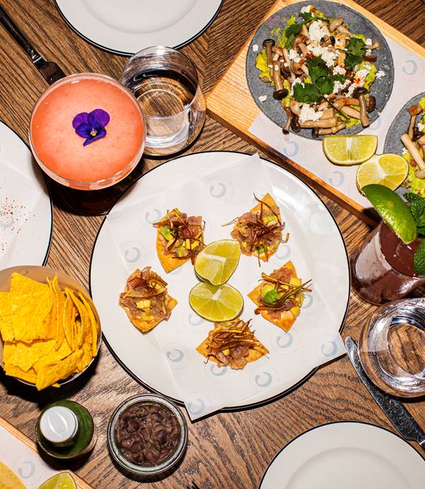 A variety of small bites, tacos, and cocktails on a table with lime wedges at Taco Tuesday at Celia in Amsterdam.