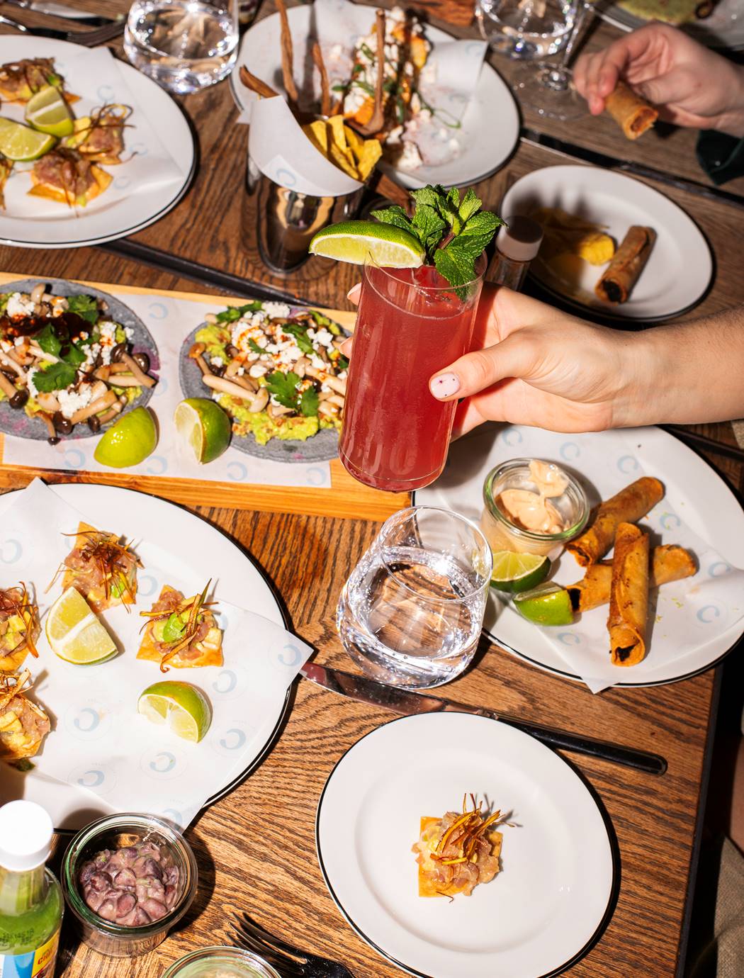 A woman holding a refreshing drink in one hand while surrounded by tacos, sauces, and crispy snacks at Taco Tuesday at Celia.