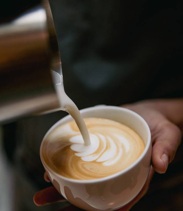 Steaming latte being poured into a white cup with intricate foam art at Celia in Amsterdam.