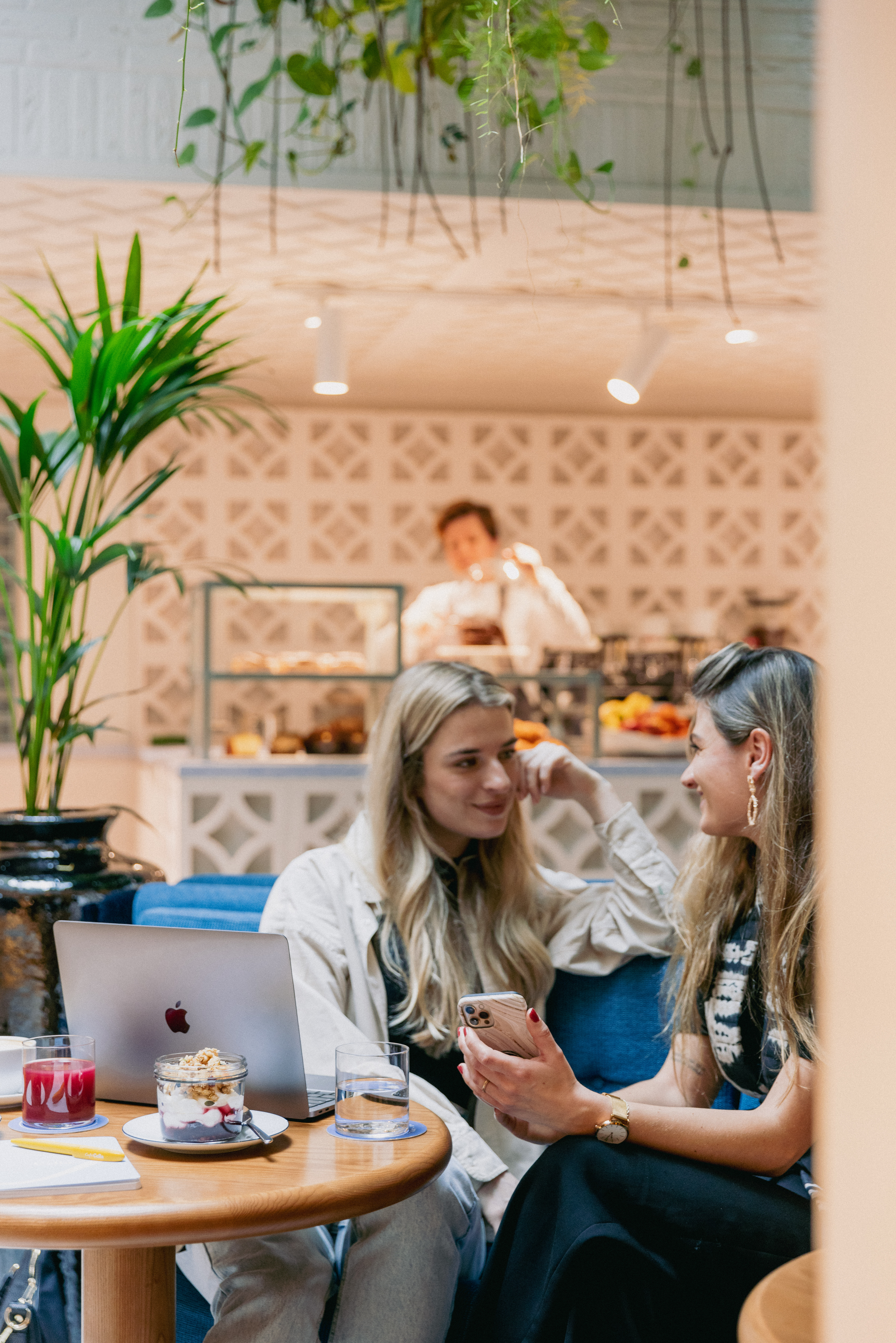 Two women sitting at a café table, laughing, with a laptop and dessert in front of them at Celia Amsterdam.