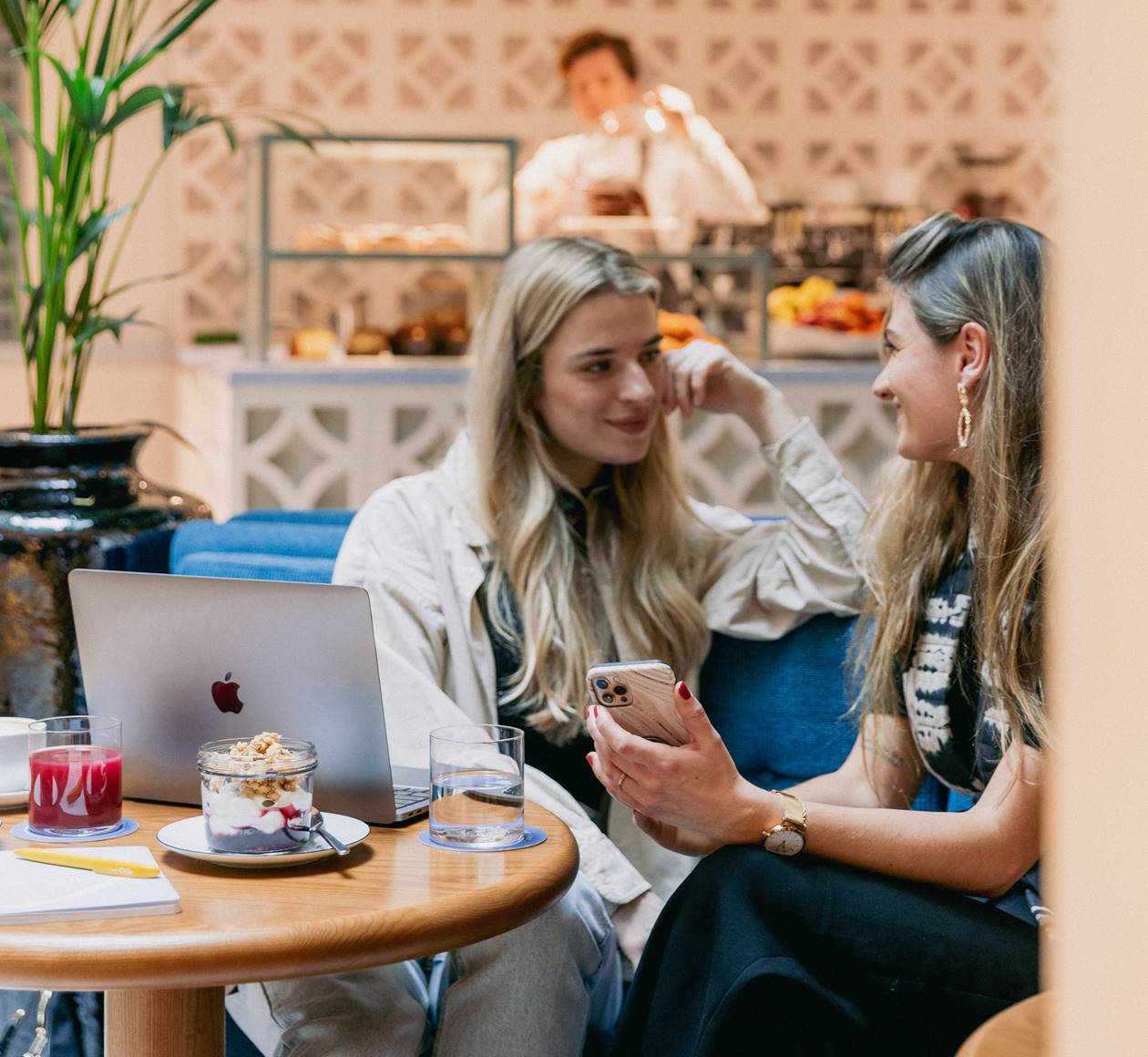 Two women sitting at a café table, laughing, with a laptop and dessert in front of them at Celia Amsterdam.
