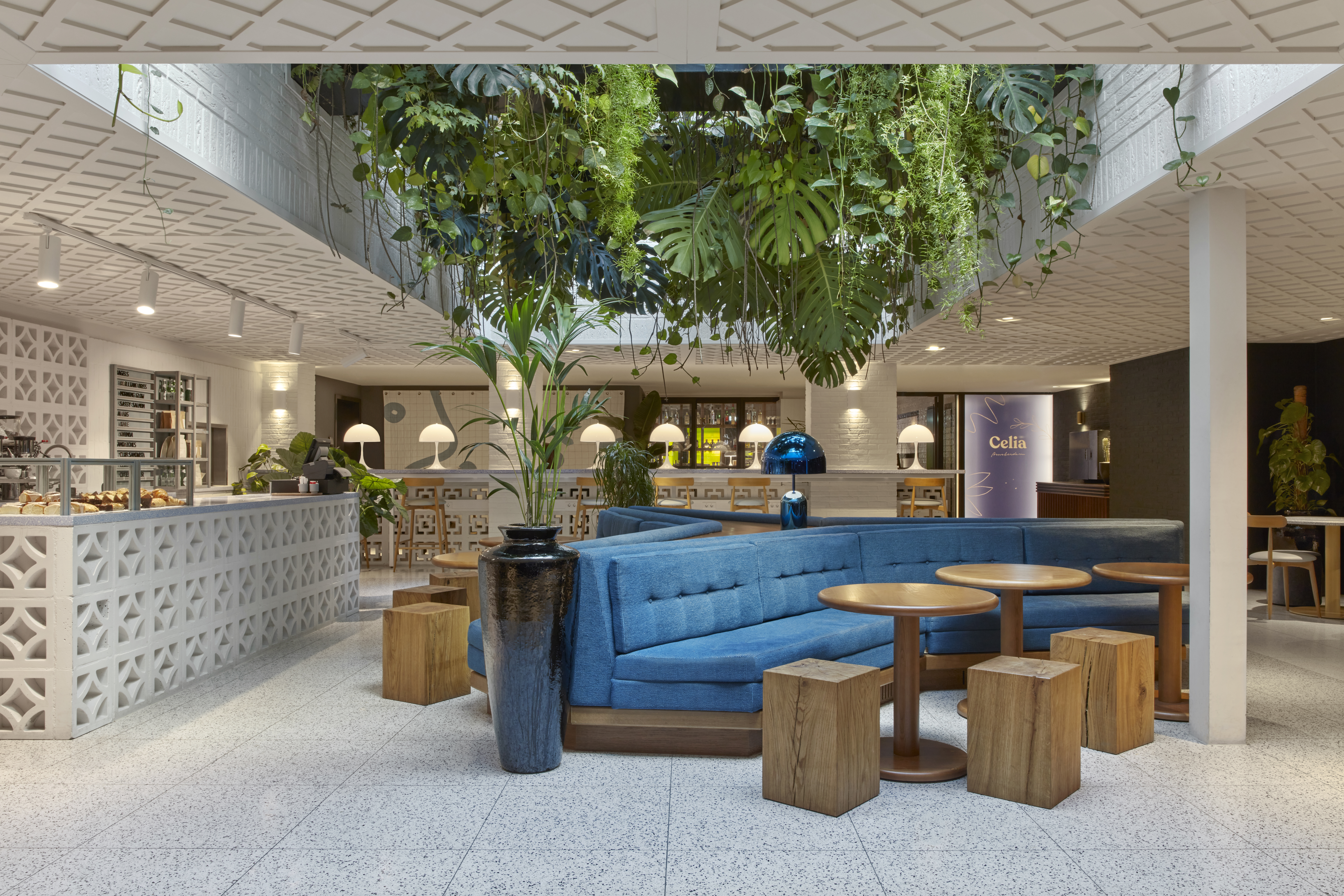 A spacious, lush café interior with blue seating, wooden tables, and hanging greenery at Celia at Kimpton De Witt.