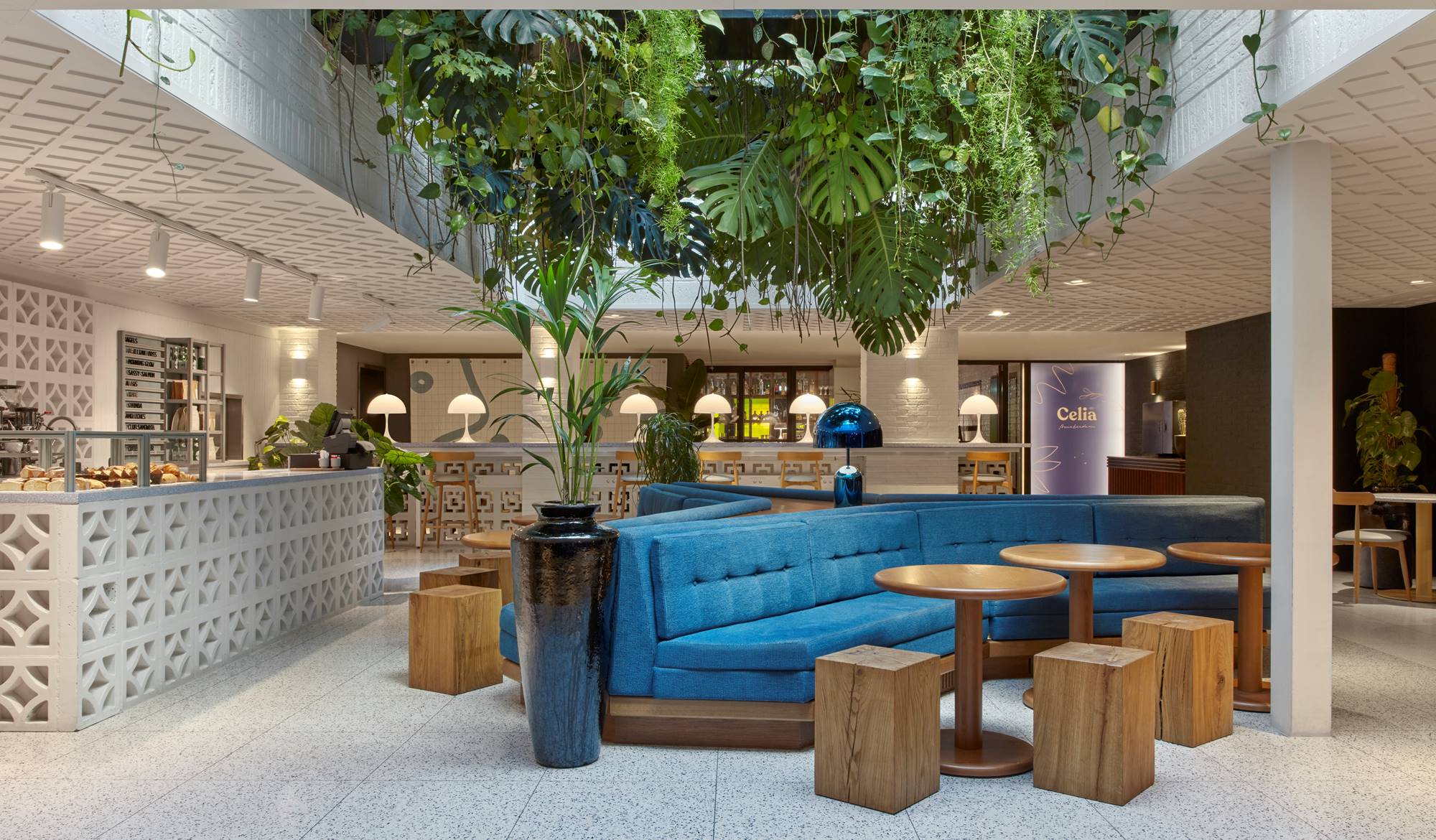 A spacious, lush café interior with blue seating, wooden tables, and hanging greenery at Celia at Kimpton De Witt.