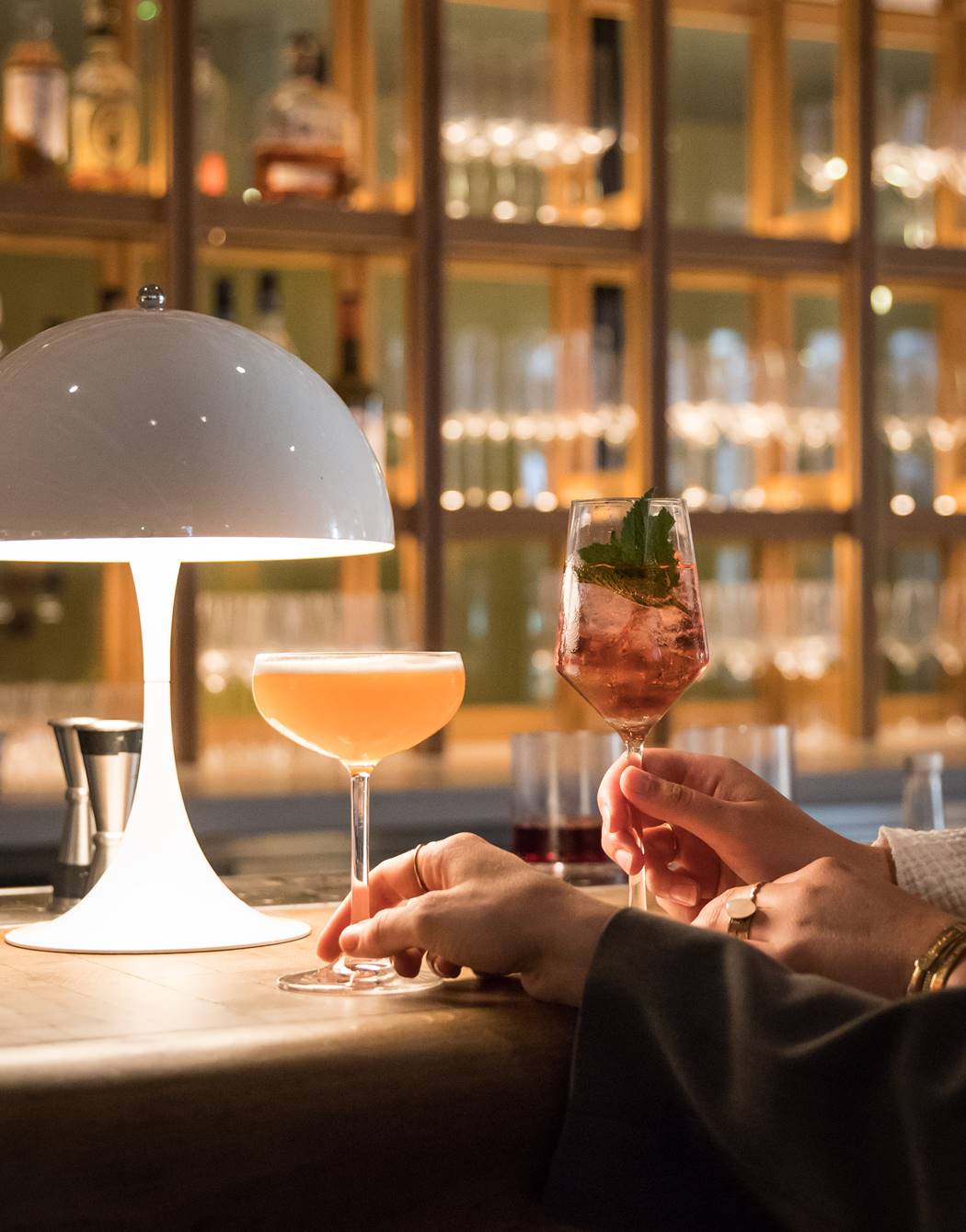 Clinking cocktail glasses in front of a well-stocked bar, with soft lighting casting a warm glow and a white lamp at Celia.