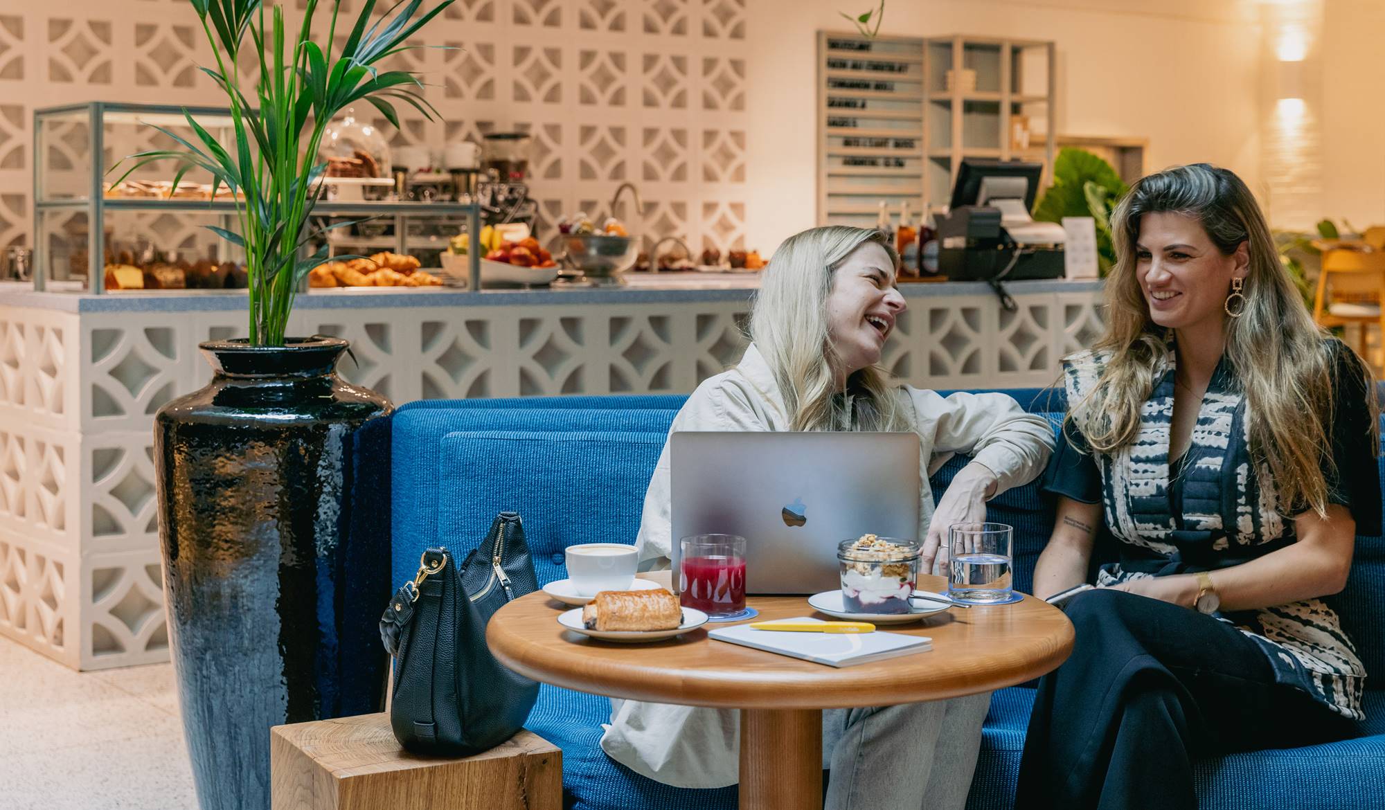 Two women laughing and enjoying drinks while sitting at a table with a laptop and food at Celia.