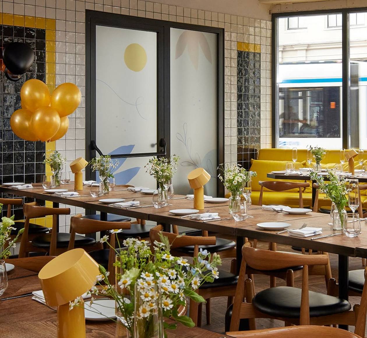 A cozy Celia restaurant setting features a long table with flowers, yellow balloons, and modern decor in Amsterdam.